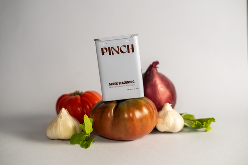 Pinch Greek seasoning tin balancing on  a red heirloom tomatoes, onions, mint leaves and garlic on a light dusty blue background