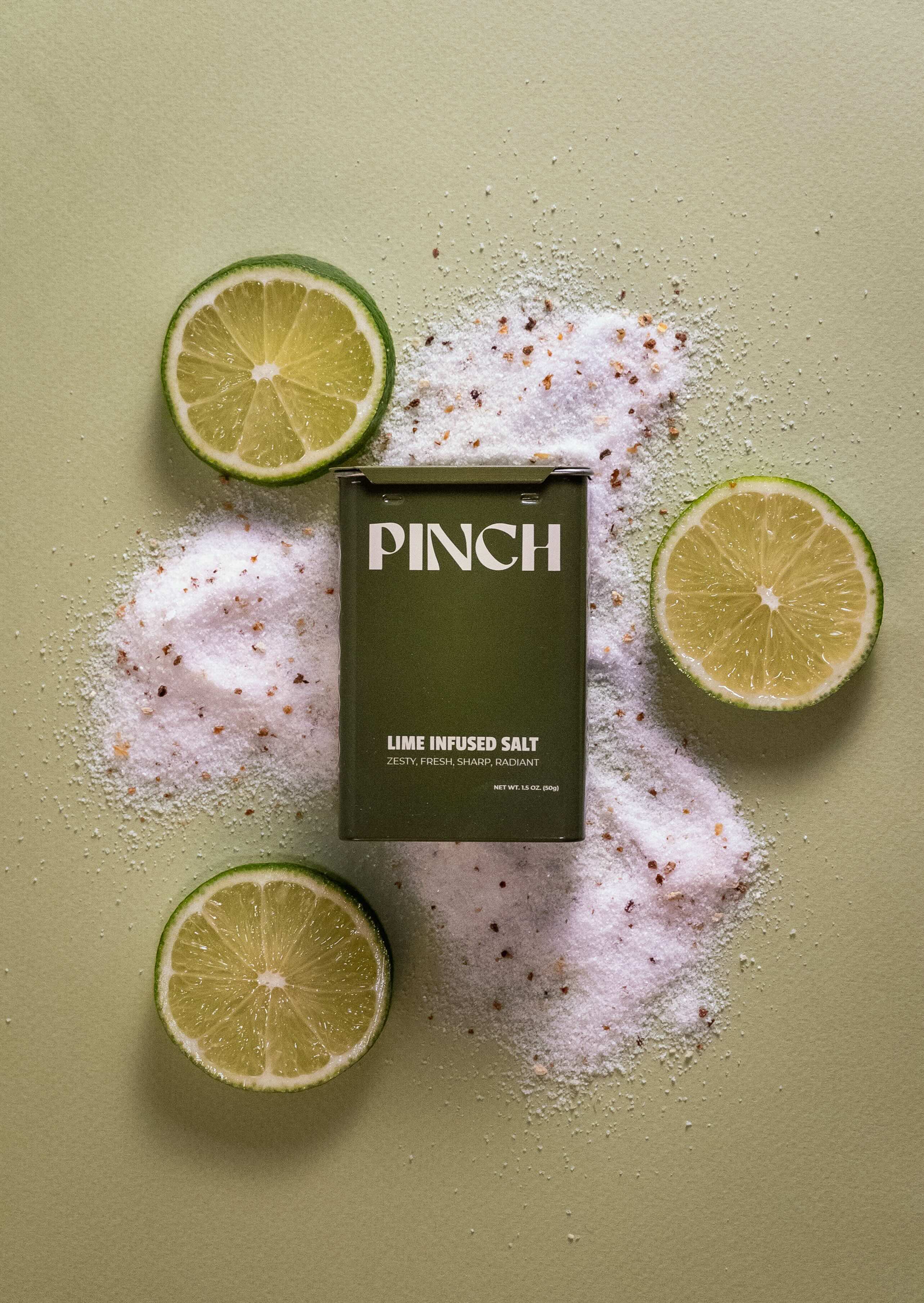 A dark olive green box of PINCH Lime Infused Salt is centered on a sage green background, surrounded by scattered lime salt salt crystals and three lime slices.