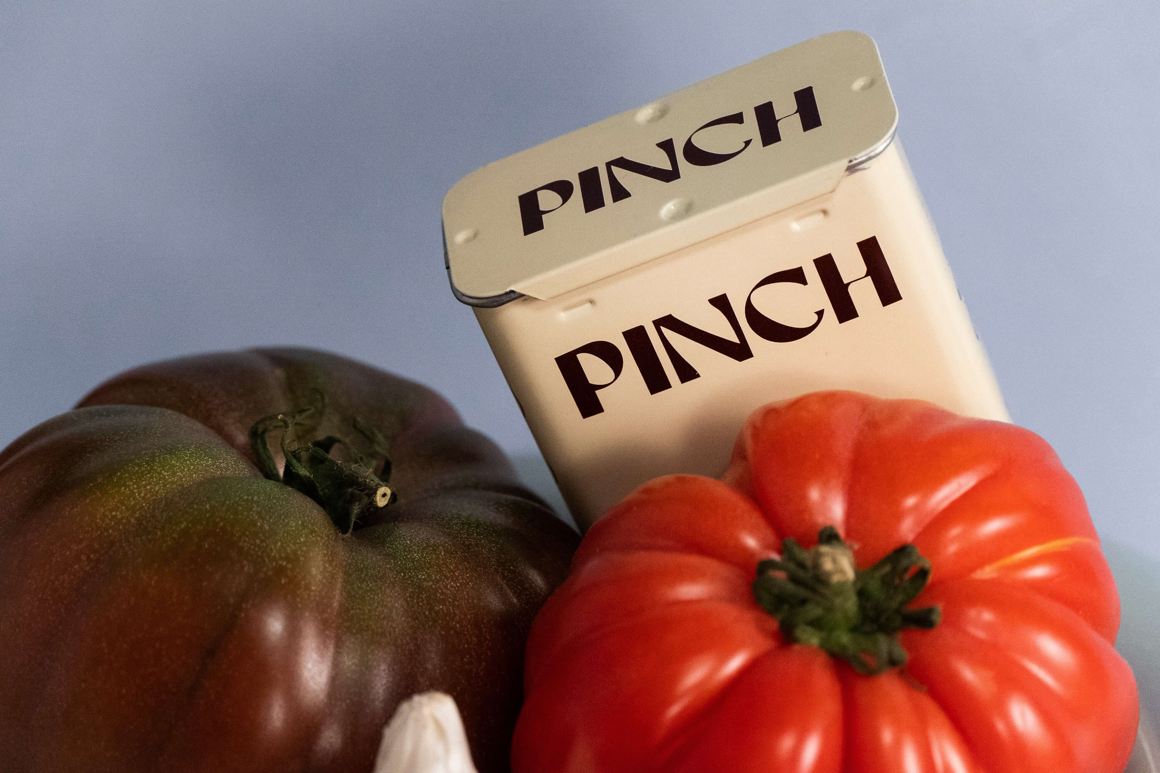 A cream-colored tin of Pinch seasoning with its lid propped open, styled with a dark heirloom tomato, a red tomato, and a garlic bulb against a soft blue-gray background.