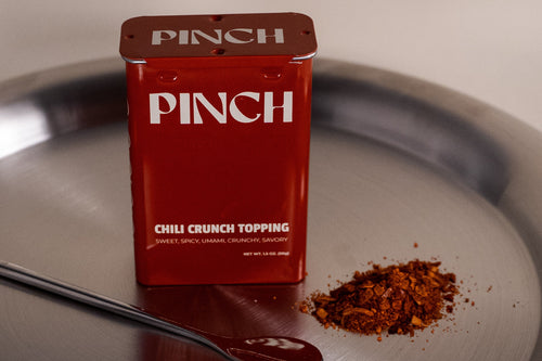 Red 'PINCH' container of chili crunch topping on a metal tray with a spoonful of topping.