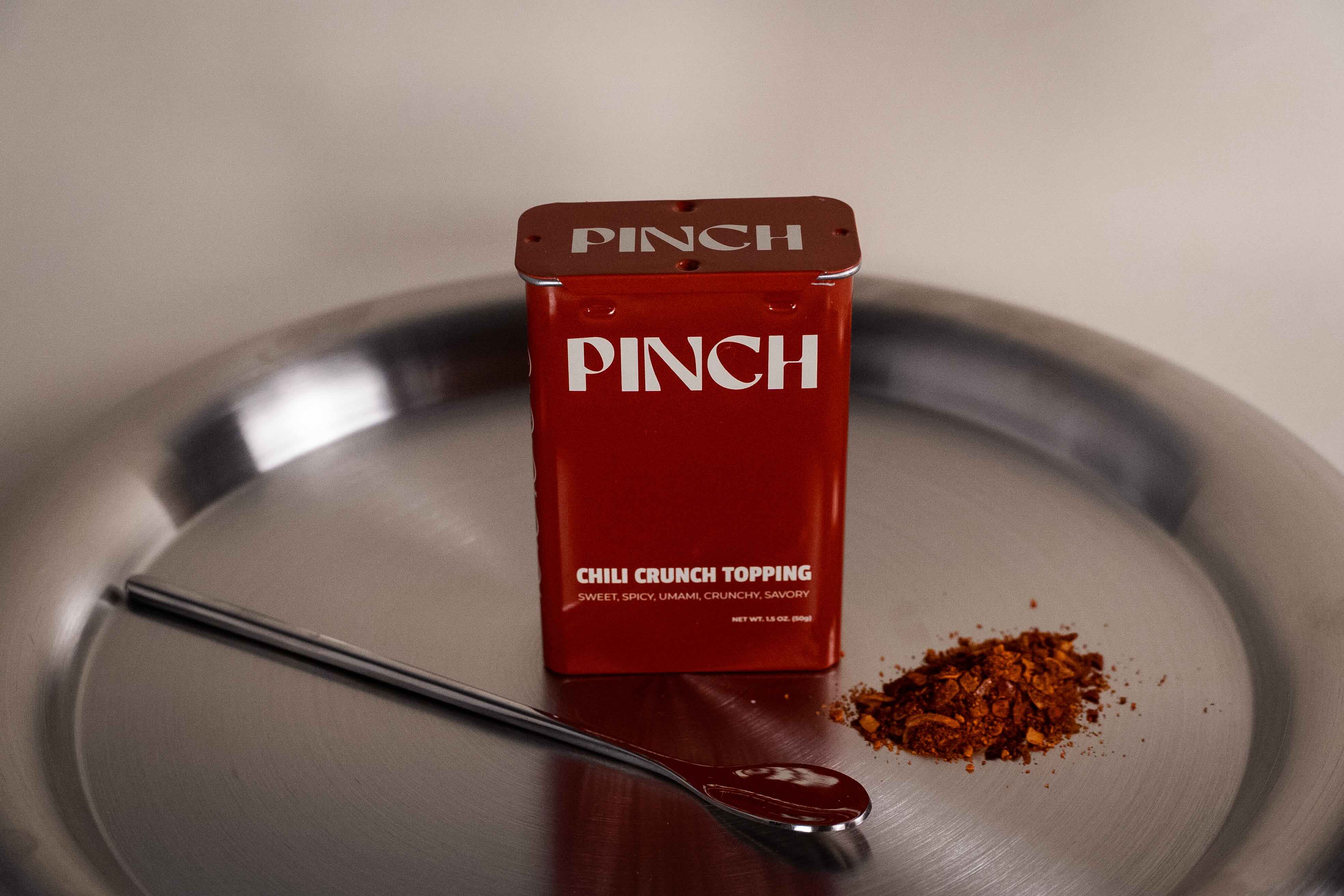 Red 'PINCH' container of chili crunch topping on a metal tray with a spoonful of topping.